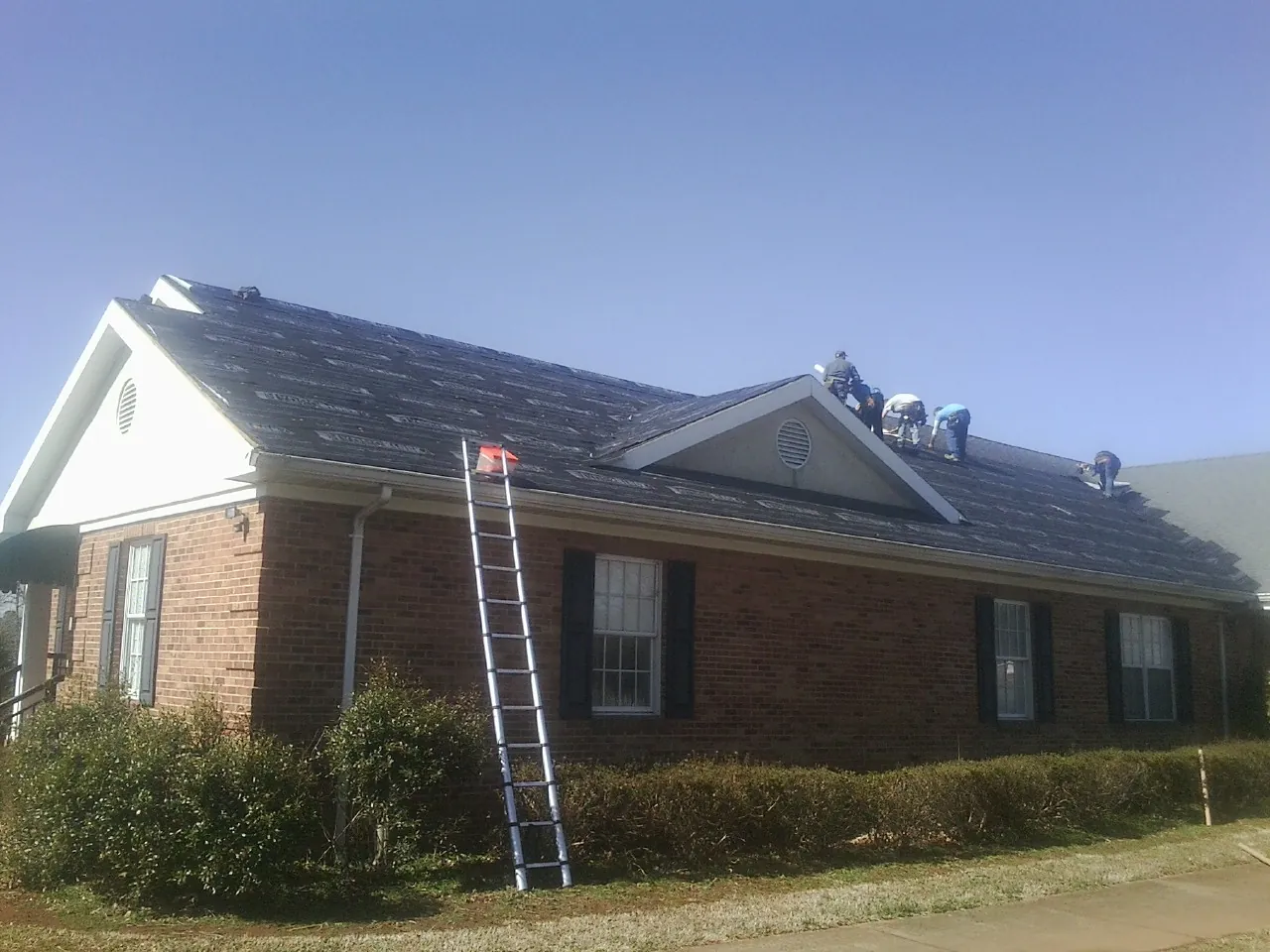 Crew tearing shingles off a brick ranch roof with a ladder set — Emergency Roof Repair work by our crew in Oriska