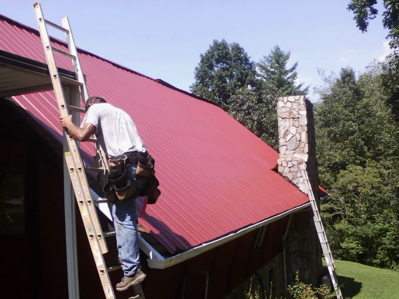 Roofer on a ladder beside a red metal roof and stone chimney — Soffit & Fascia Repair by Roofing Brothers in Oriska